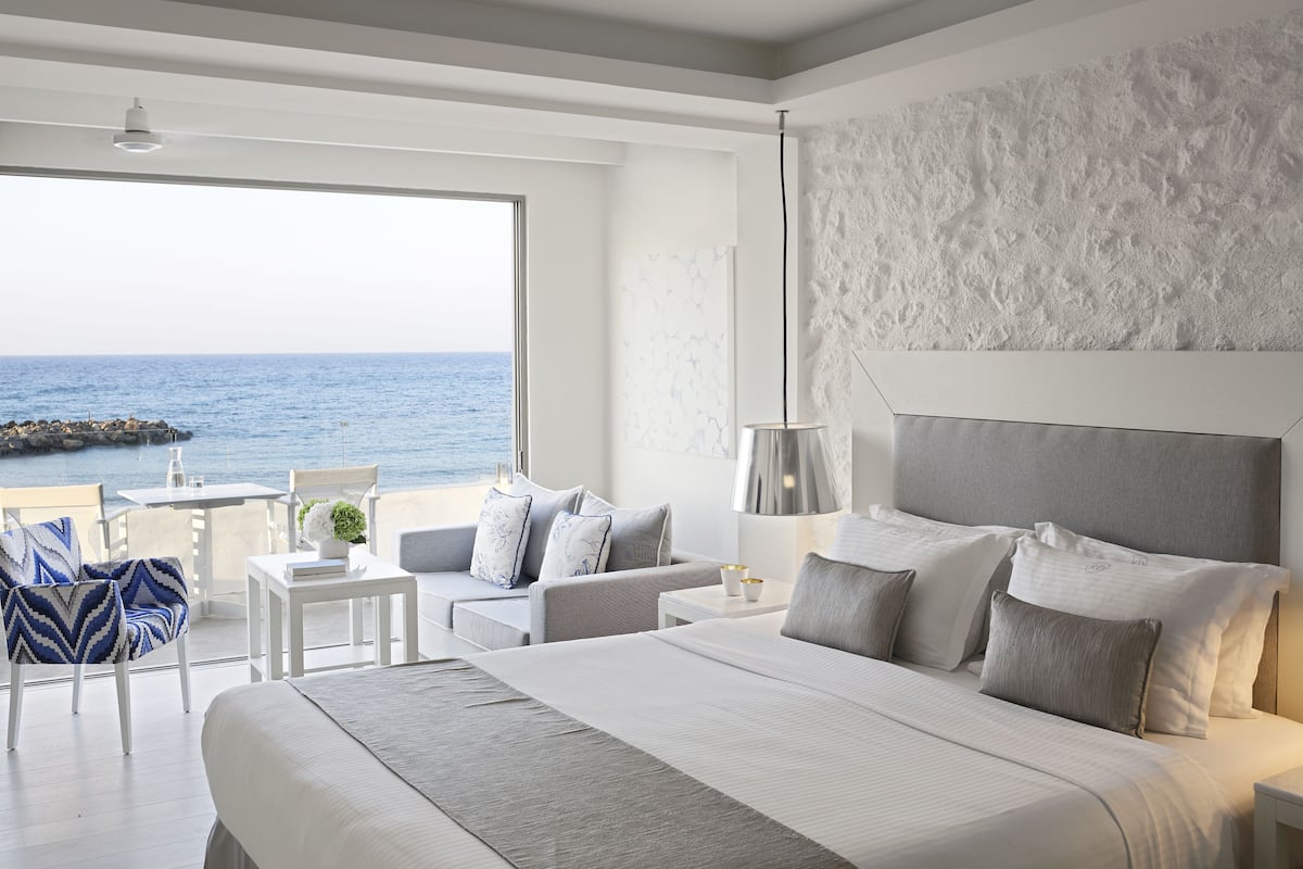 Ein modernes Hotelzimmer mit einem großen Bett, einem Balkon mit Meerblick und einer Essecke mit Tisch und Stühlen.
