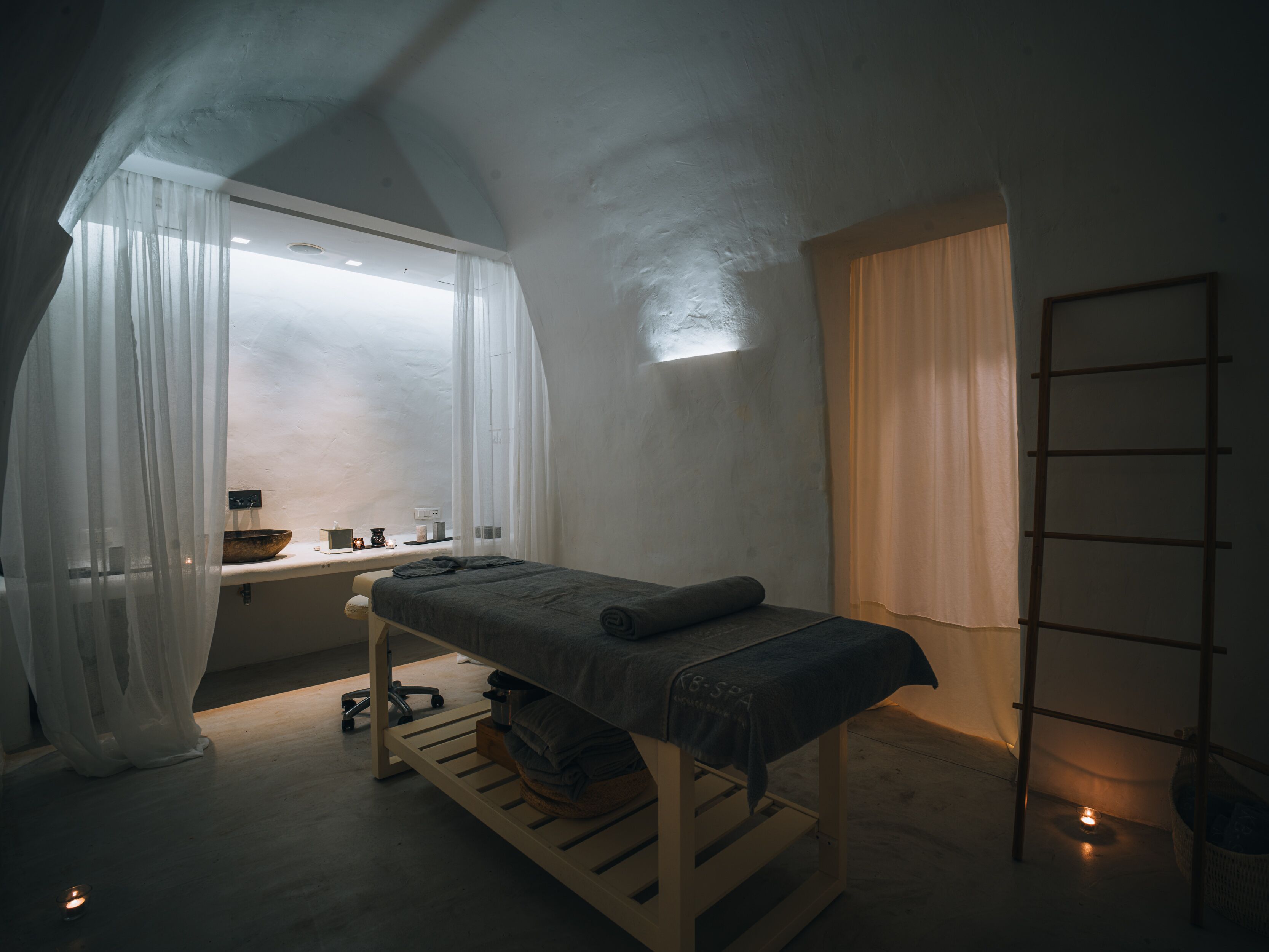 A minimalist bedroom with a single bed, a bedside table, a window with sheer curtains, and a ladder against the wall.