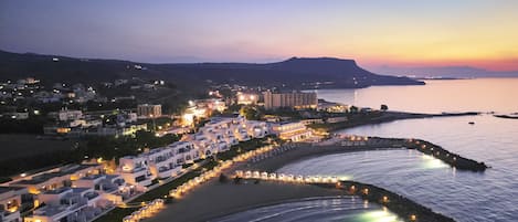 A coastal town at dusk with illuminated buildings, a beach, and a calm sea.