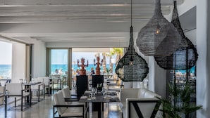 A modern dining area with a view of the sea, featuring black woven pendant lights and white furniture.