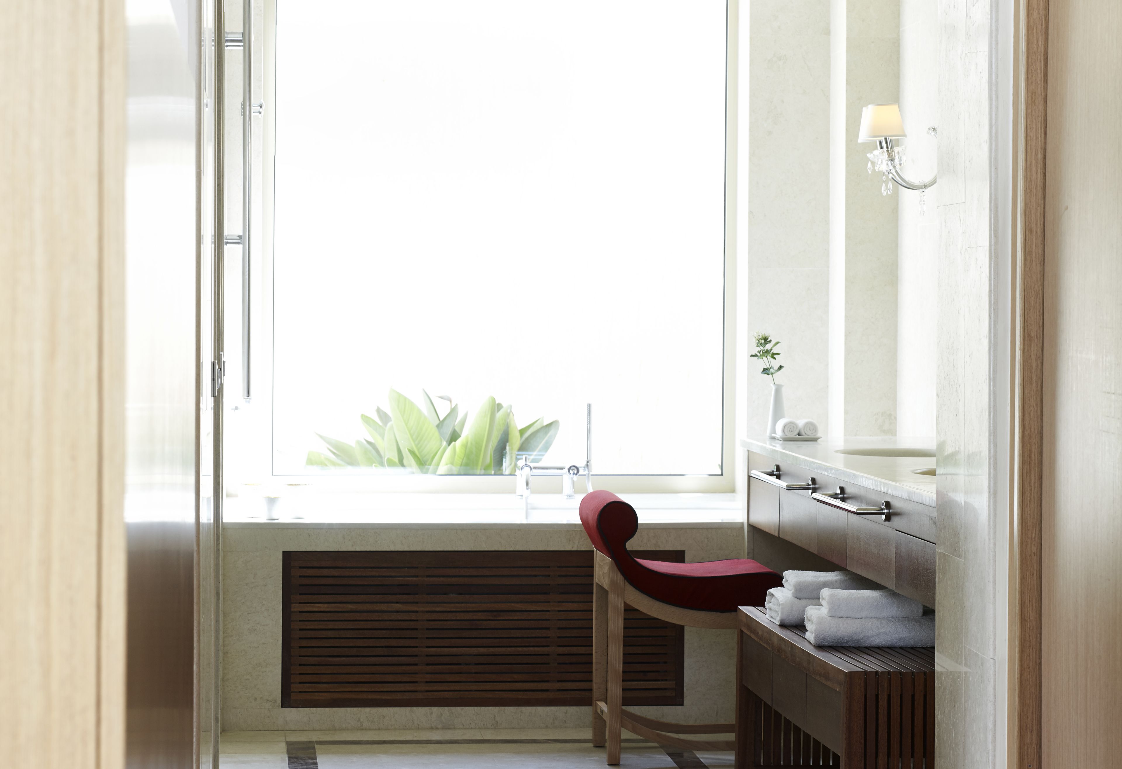 A bathroom with a wooden vanity, a red chair, a window with a view of greenery, and a wall-mounted light fixture.