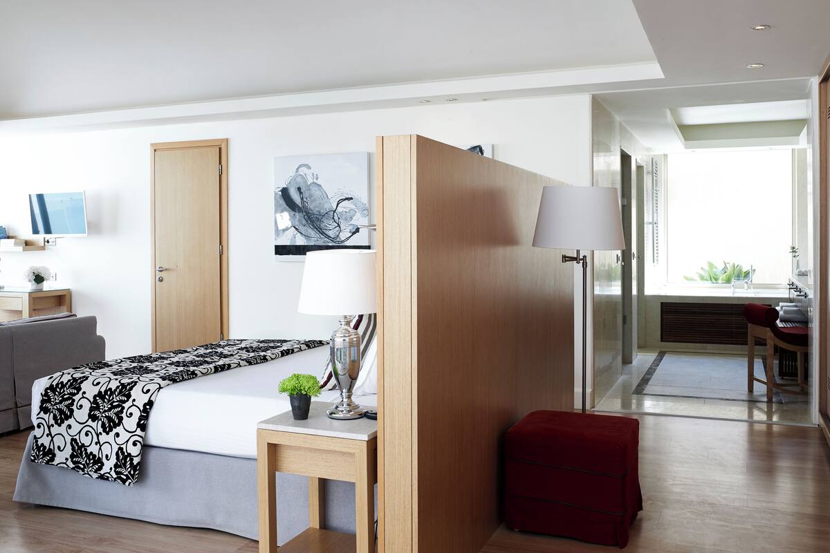 Ein modernes Hotelzimmer mit Bett, Nachttisch, Lampe und einem roten Hocker. Es gibt einen Schreibtisch mit Stuhl und einen Fernseher an der Wand.