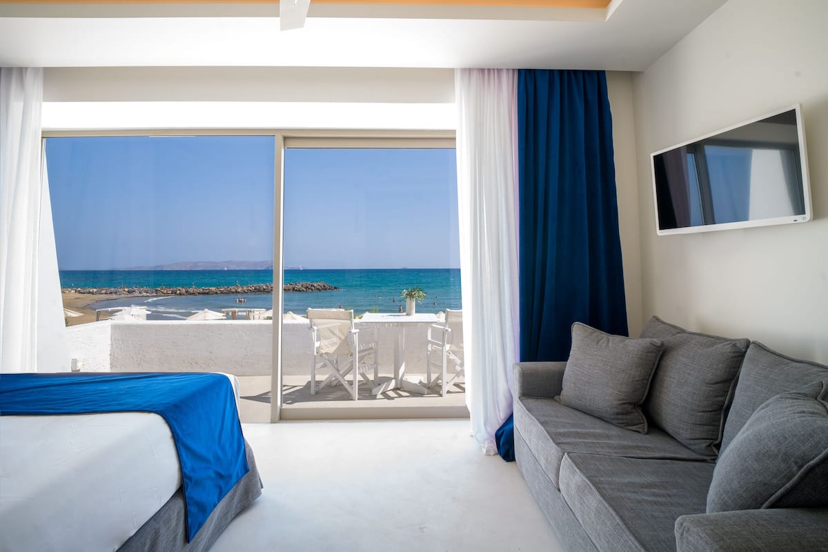 Ein Hotelzimmer mit einem Bett, einem grauen Sofa, einem Fernseher und einem Balkon mit Meerblick.