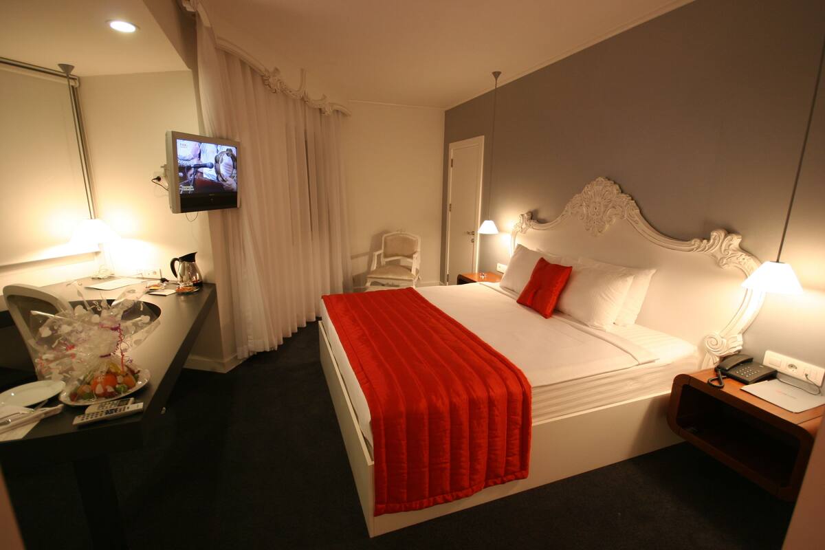 A hotel room with a bed, a desk with a chair, a television, and a nightstand with a lamp.