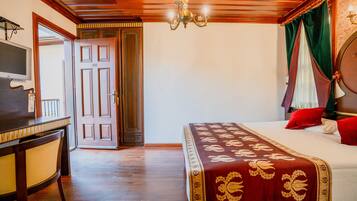 A hotel room with a wooden ceiling, a bed with a red and gold bedspread, a desk with a chair, and a television on the wall.