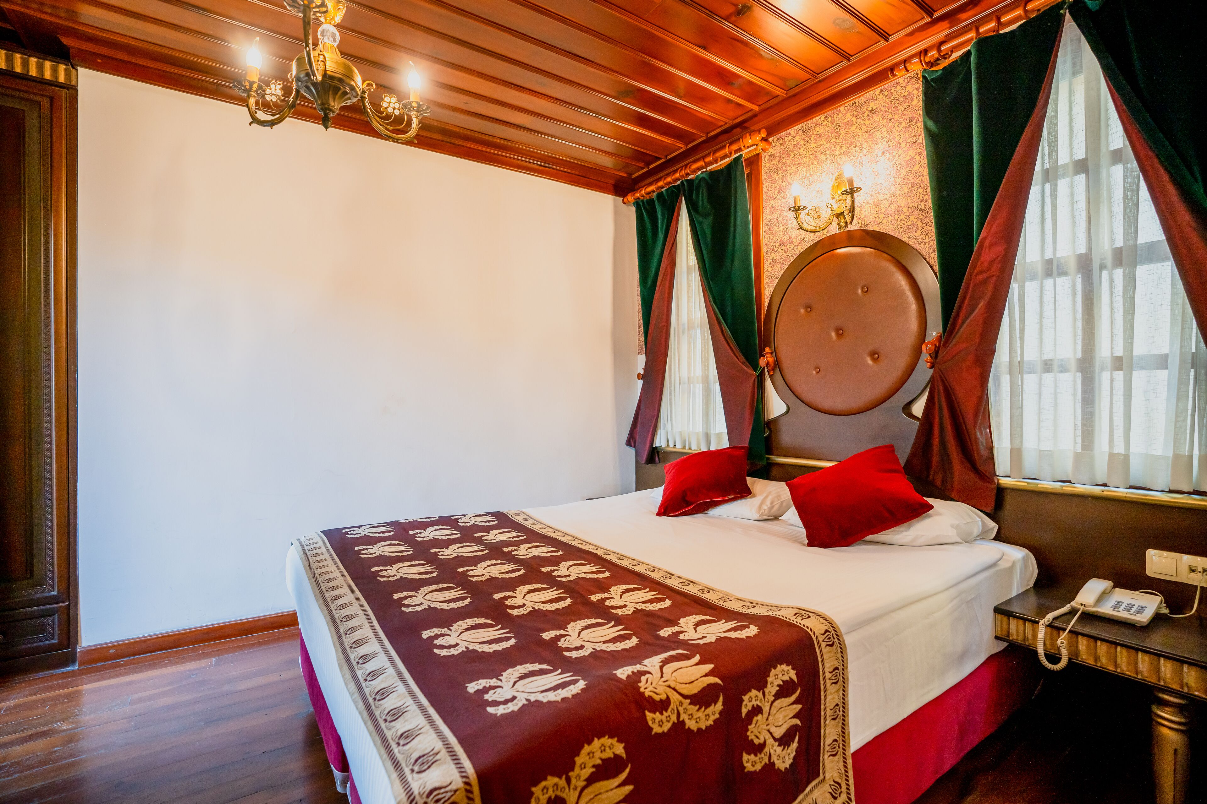 Een hotelkamer met een houten plafond, een bed met een rood-witte dekbedovertrek, een ronde spiegel boven het bed en een bedtafeltelefoon.