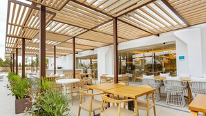An outdoor cafe area with wooden tables and chairs, potted plants, and a glass wall overlooking the interior.
