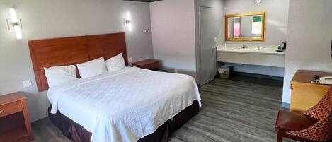 A hotel room with a large bed, a wooden headboard, a bedside table, a chair, a desk, a mirror, and a bathroom with a sink and toilet.