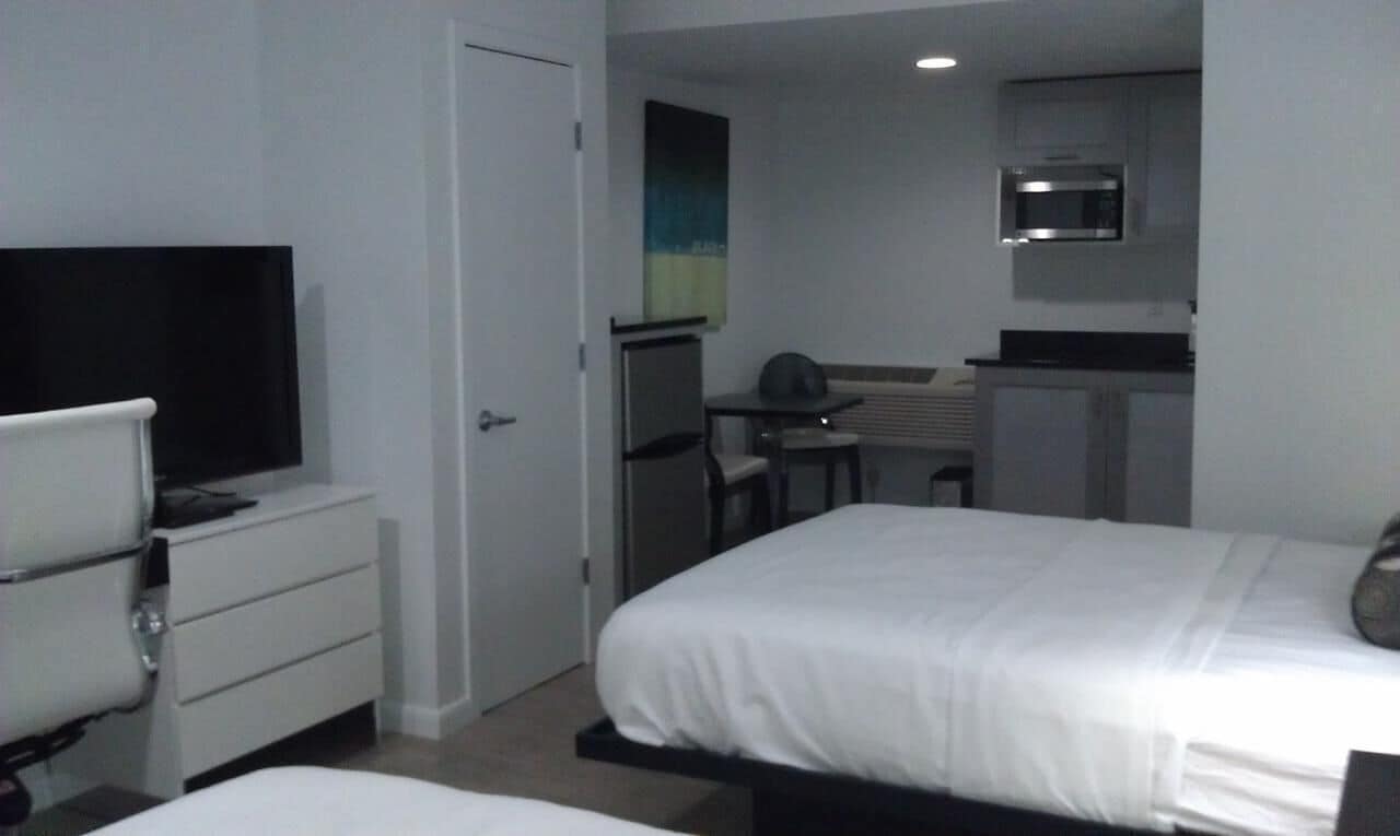Habitación de hotel con cama, escritorio, silla, televisión y kitchenette.