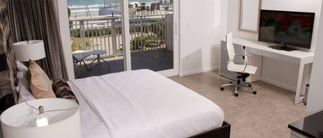 A hotel room with a bed, a desk with a computer, a TV, and a balcony with a view of the beach.