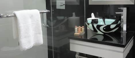 Un baño moderno con ducha envuelta en vidrio, un lavamanos negro con un recipiente de cristal y un espejo grande sobre el lavamanos.