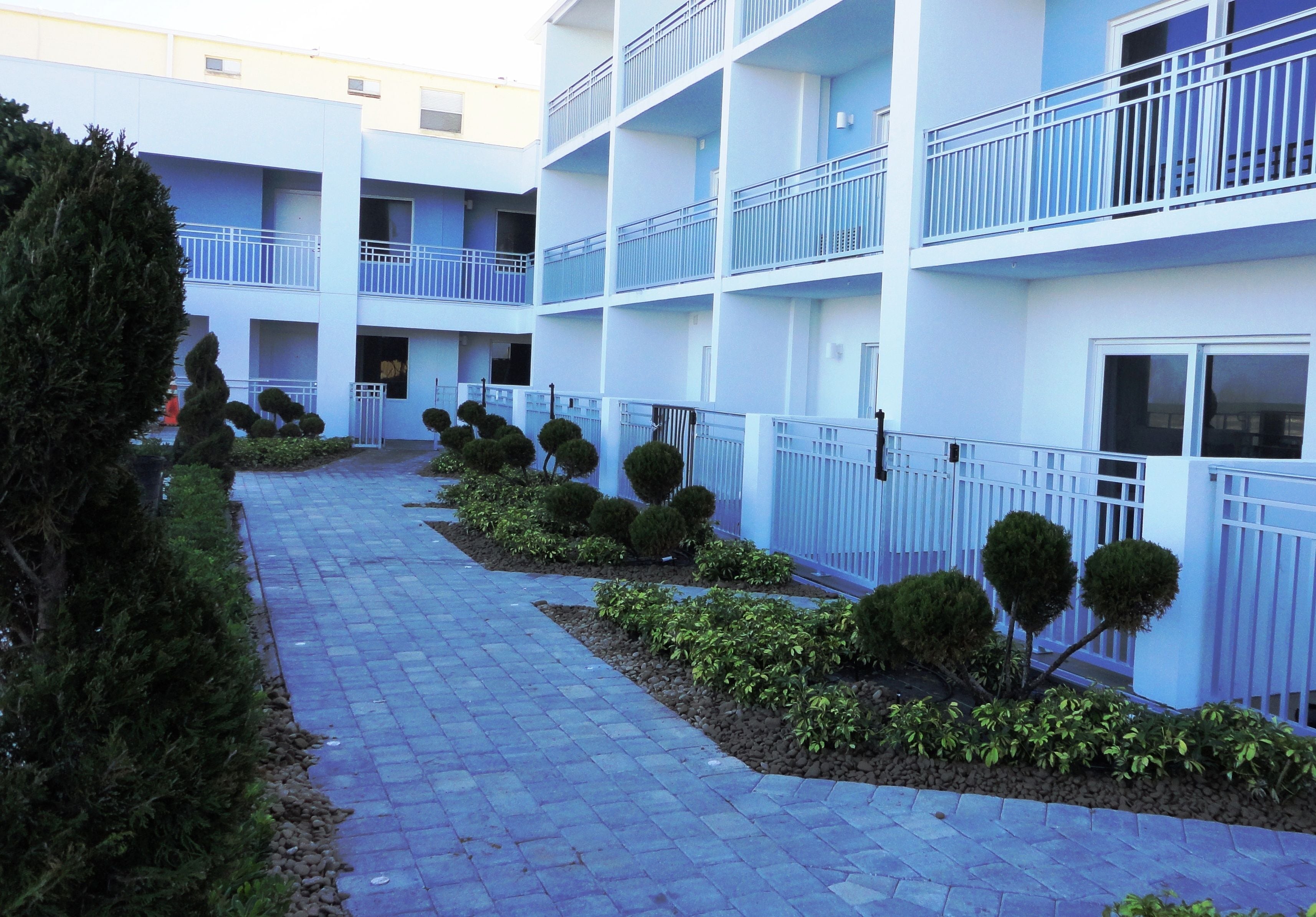 Un hotel de varios pisos con fachada azul y blanca, un sendero pavimentado y setos recortados a lo largo del camino.