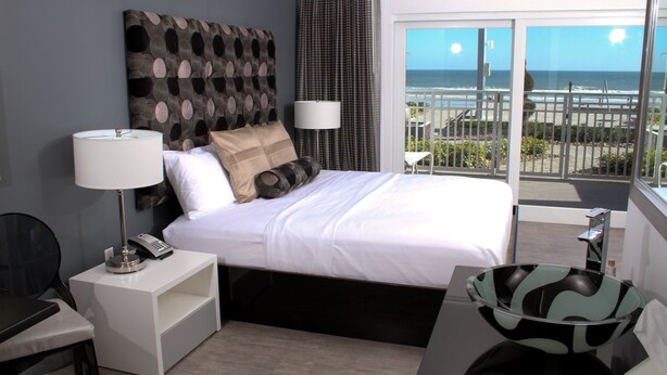 A modern hotel room with a large bed, a desk, a chair, a nightstand with a lamp, and a view of the beach through the balcony. - Lotus Boutique Inn & Suites (Ormond Beach)