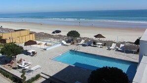 A pool area with lounge chairs and umbrellas, overlooking a beach with people and a car. - Lotus Boutique Inn & Suites (Ormond Beach)