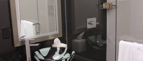 Een moderne badkamer met een douche met glazen wand, een glazen wastafel met een zwart aanrechtblad en een spiegel boven de wastafel.