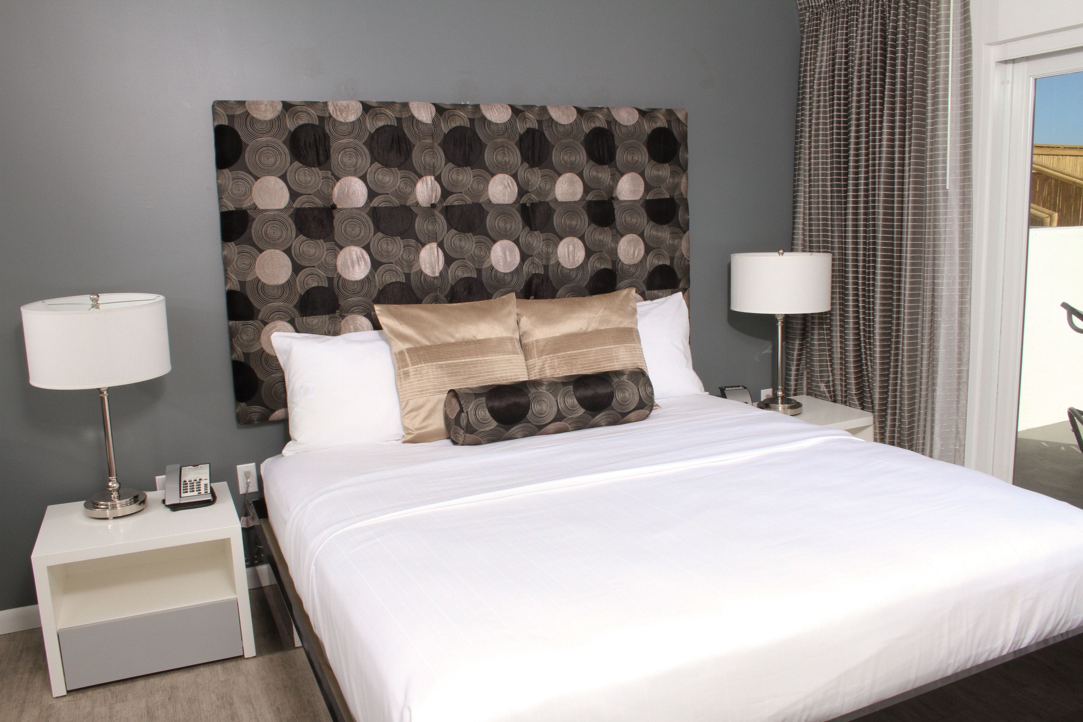 A neatly made bed with white linens and decorative pillows, a bedside lamp, and a small nightstand with a phone and a lamp.