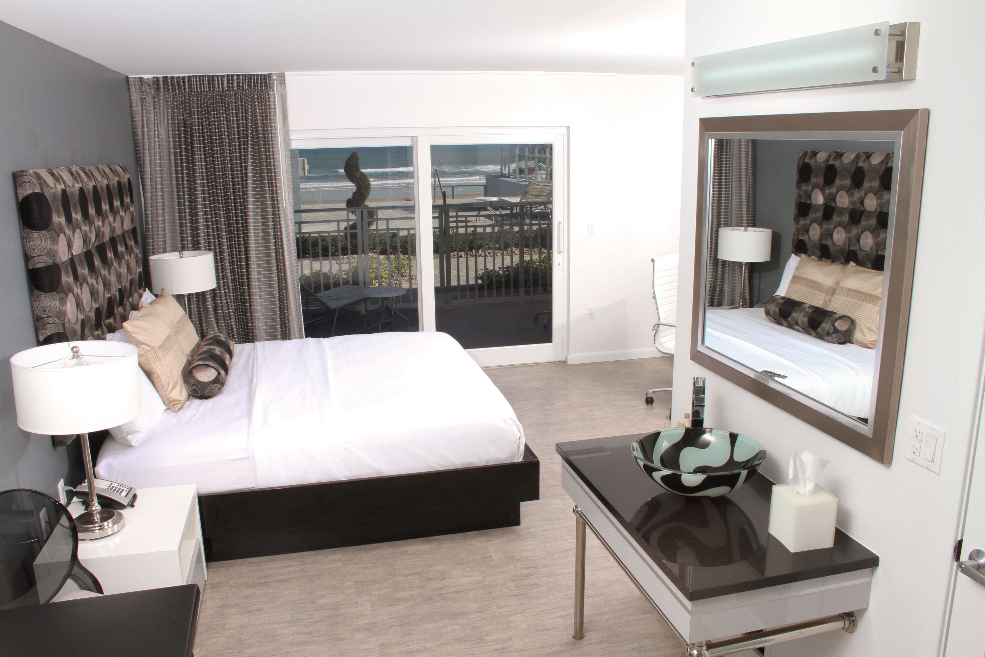 A modern hotel room with a large bed, a desk with a bowl and tissues, a mirror, and a view of the beach through the sliding doors.