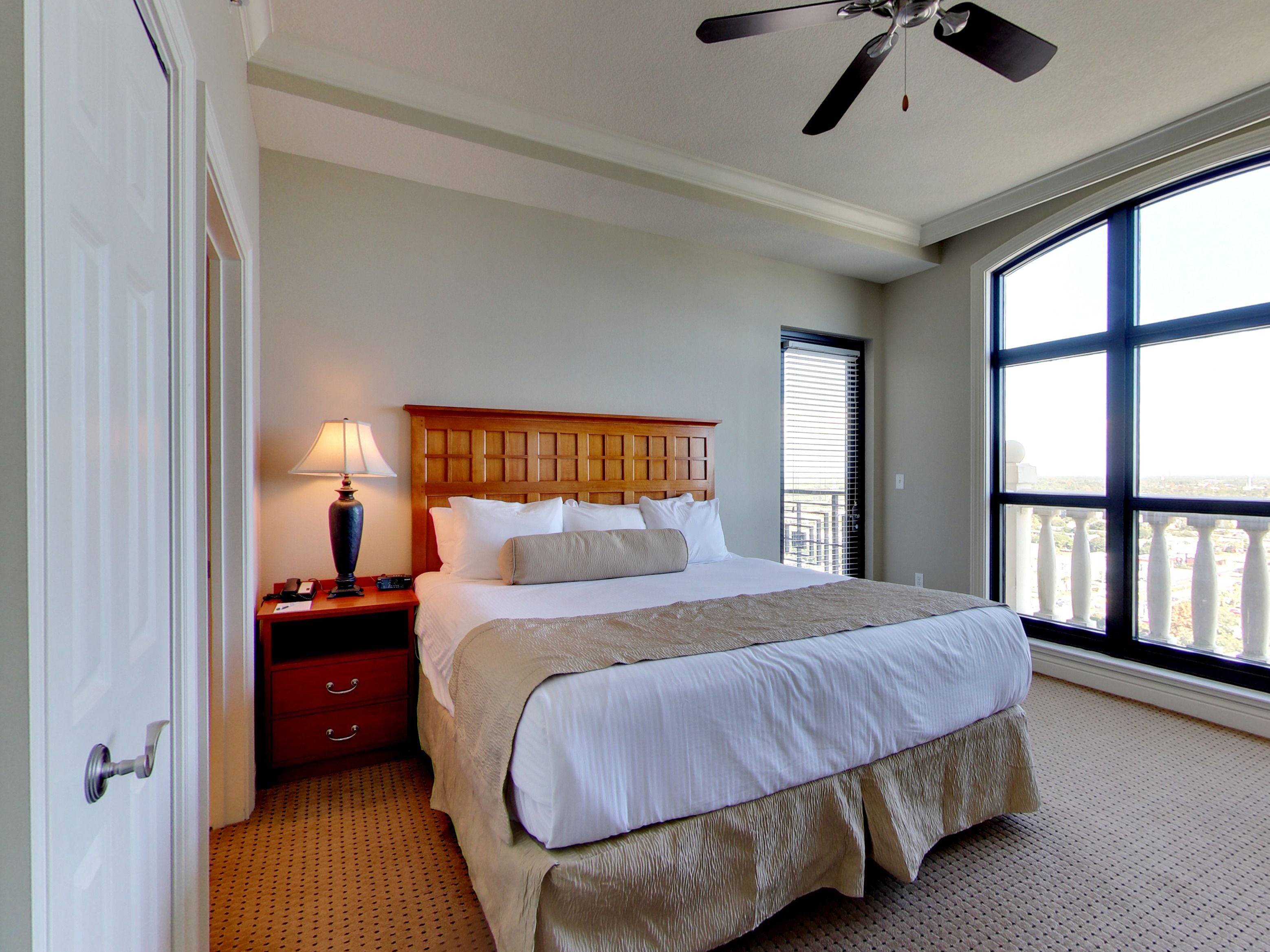 A hotel room with a large bed, a nightstand, a lamp, a ceiling fan, and a large window with a view.