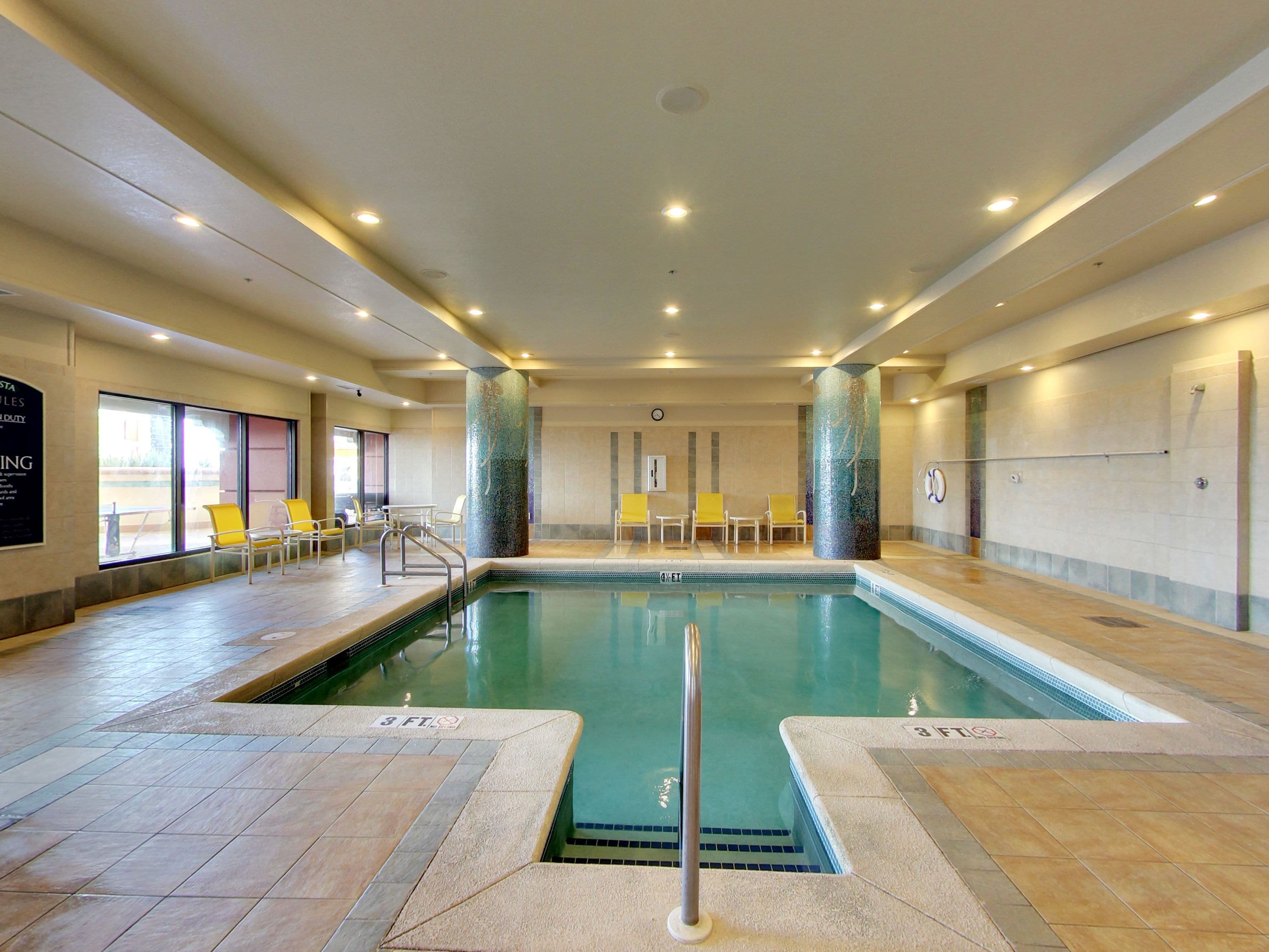 An indoor swimming pool with clear water, tiled walls, and a few yellow chairs around the pool area.