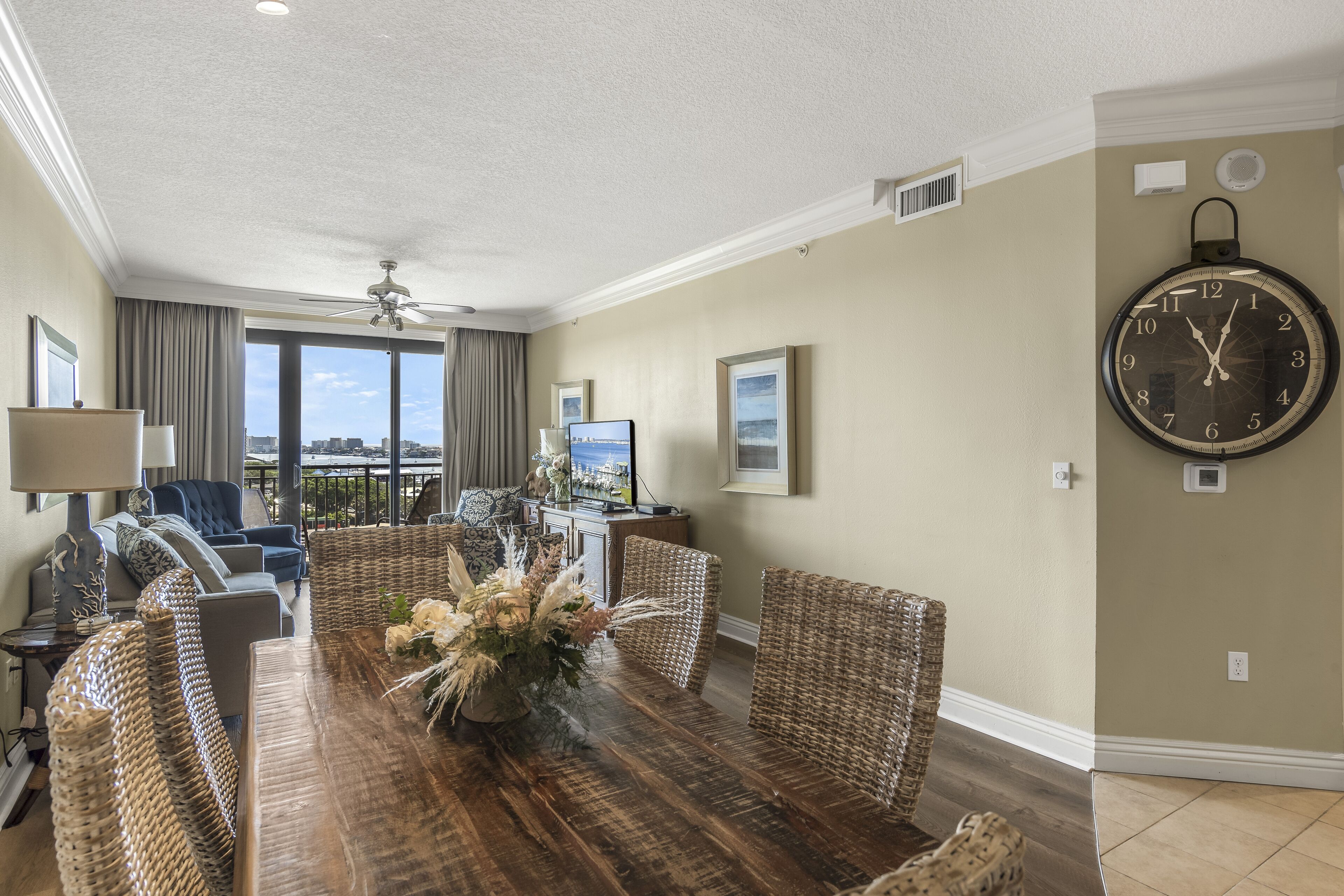 A spacious living room with a large dining table, wicker furniture, and a large wall clock.