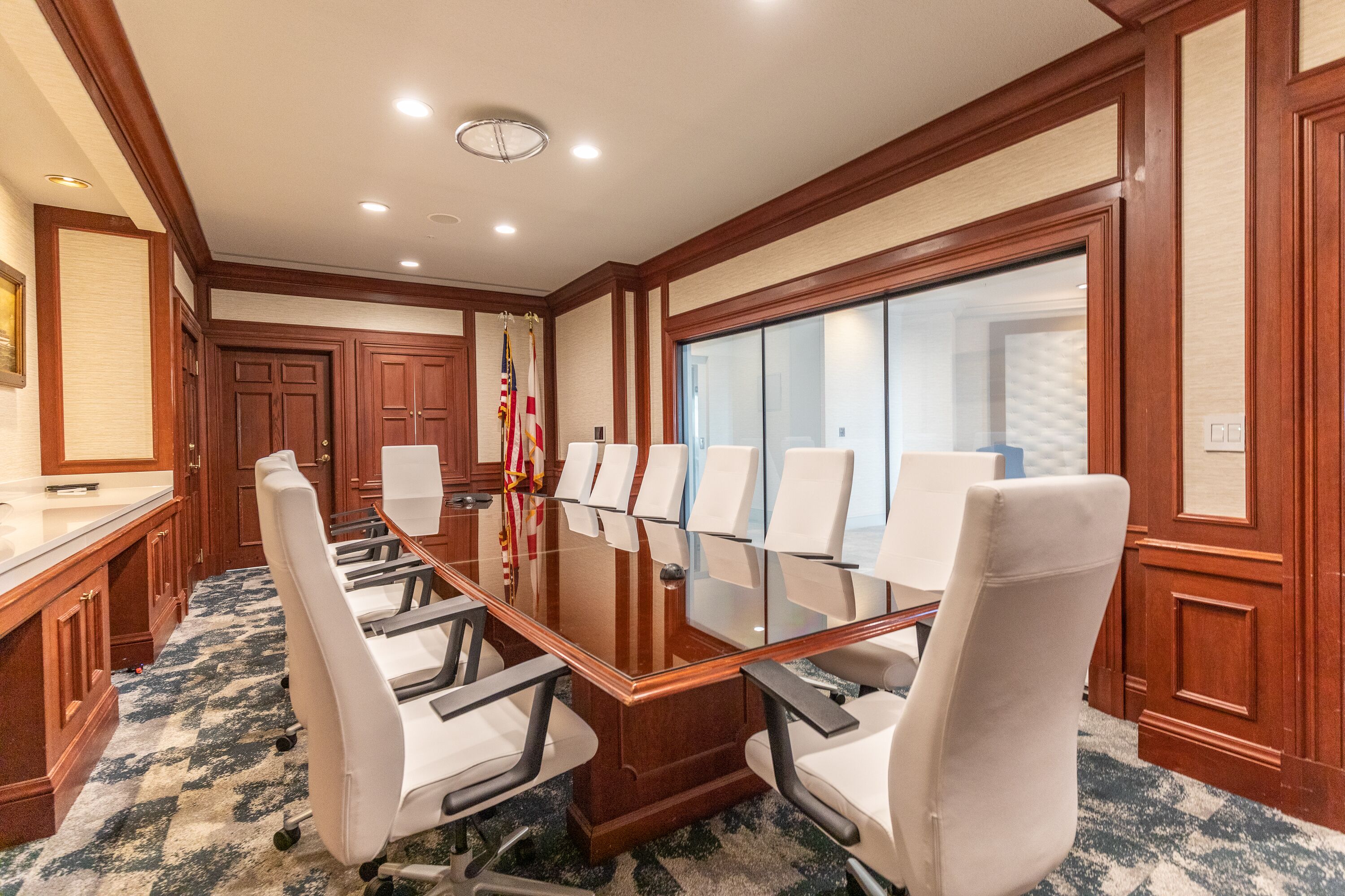 A conference room with a long table, white chairs, wooden paneling, and a large window.