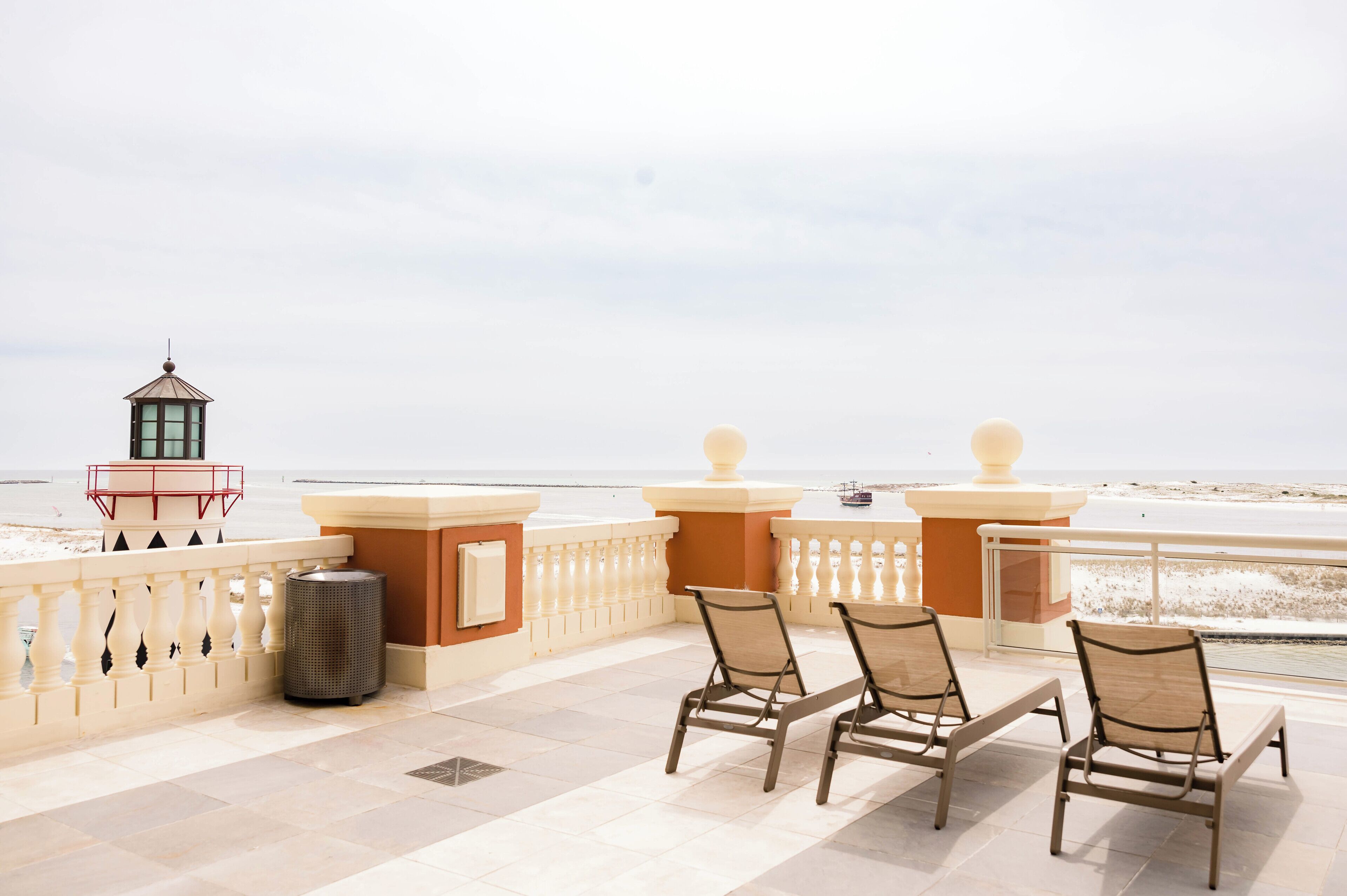 Ein Balkon mit drei Korbstühlen, einem Mülleimer und einer kleinen roten und weißen Konstruktion mit Blick auf einen Strand.