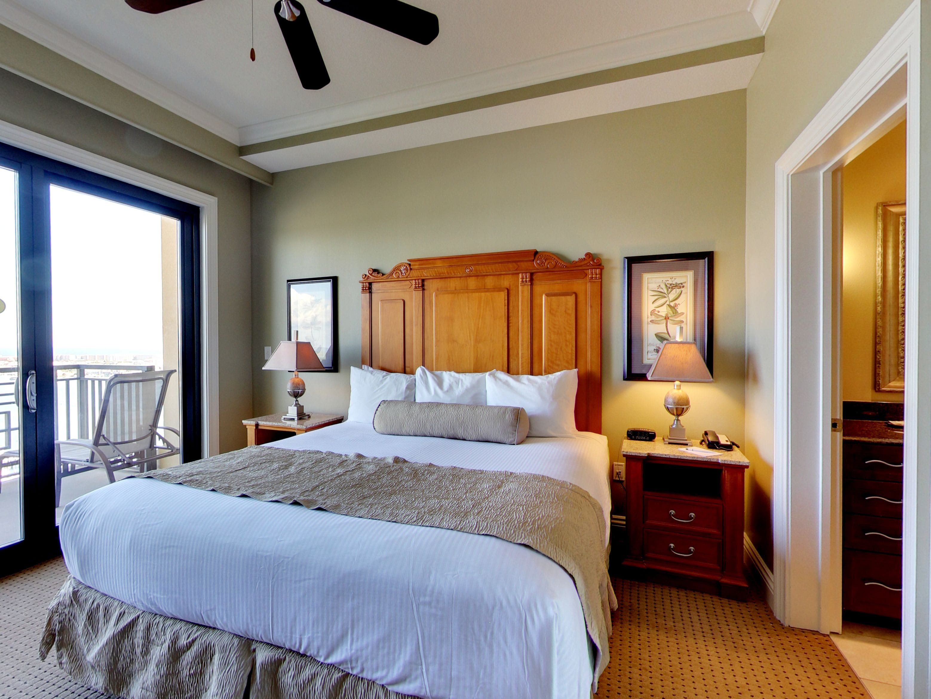 A hotel room with a large bed, a wooden headboard, a nightstand with a lamp, a chair by the window, and a door leading to a balcony.