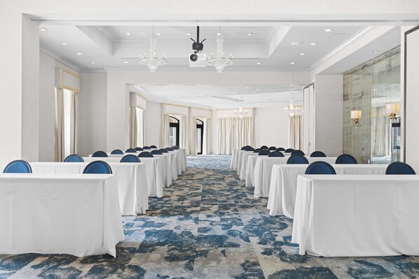 Un salón de conferencias con filas de mesas y sillas, una alfombra estampada y candelabros.