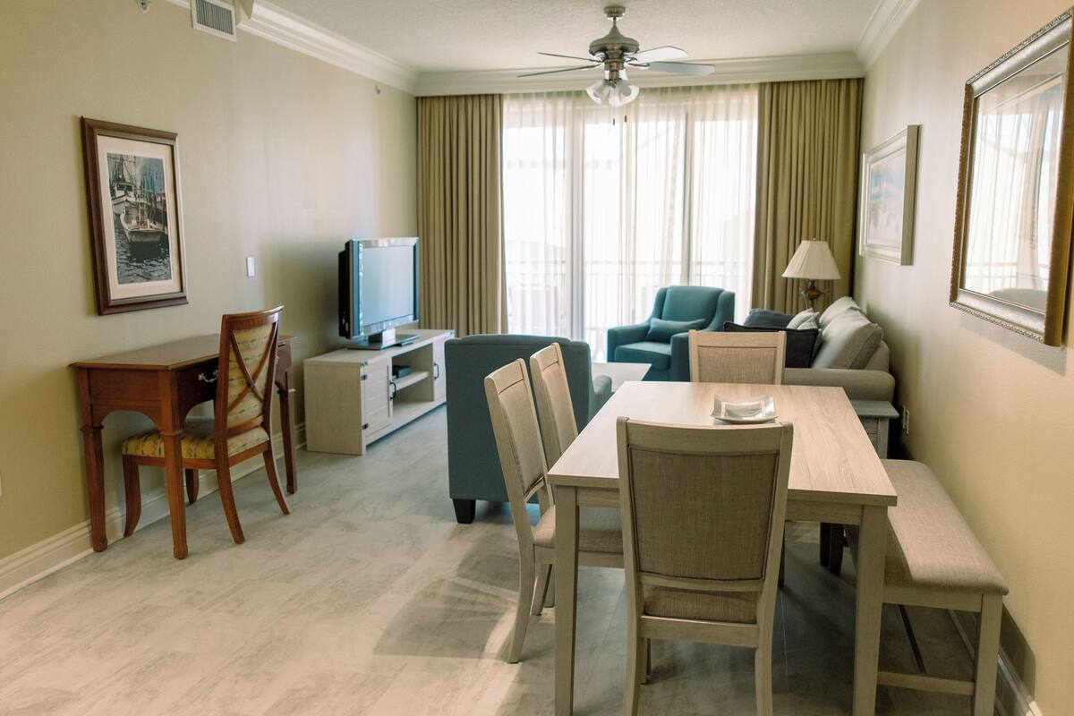 A hotel room with a dining area, a TV, a ceiling fan, and a large window with curtains.