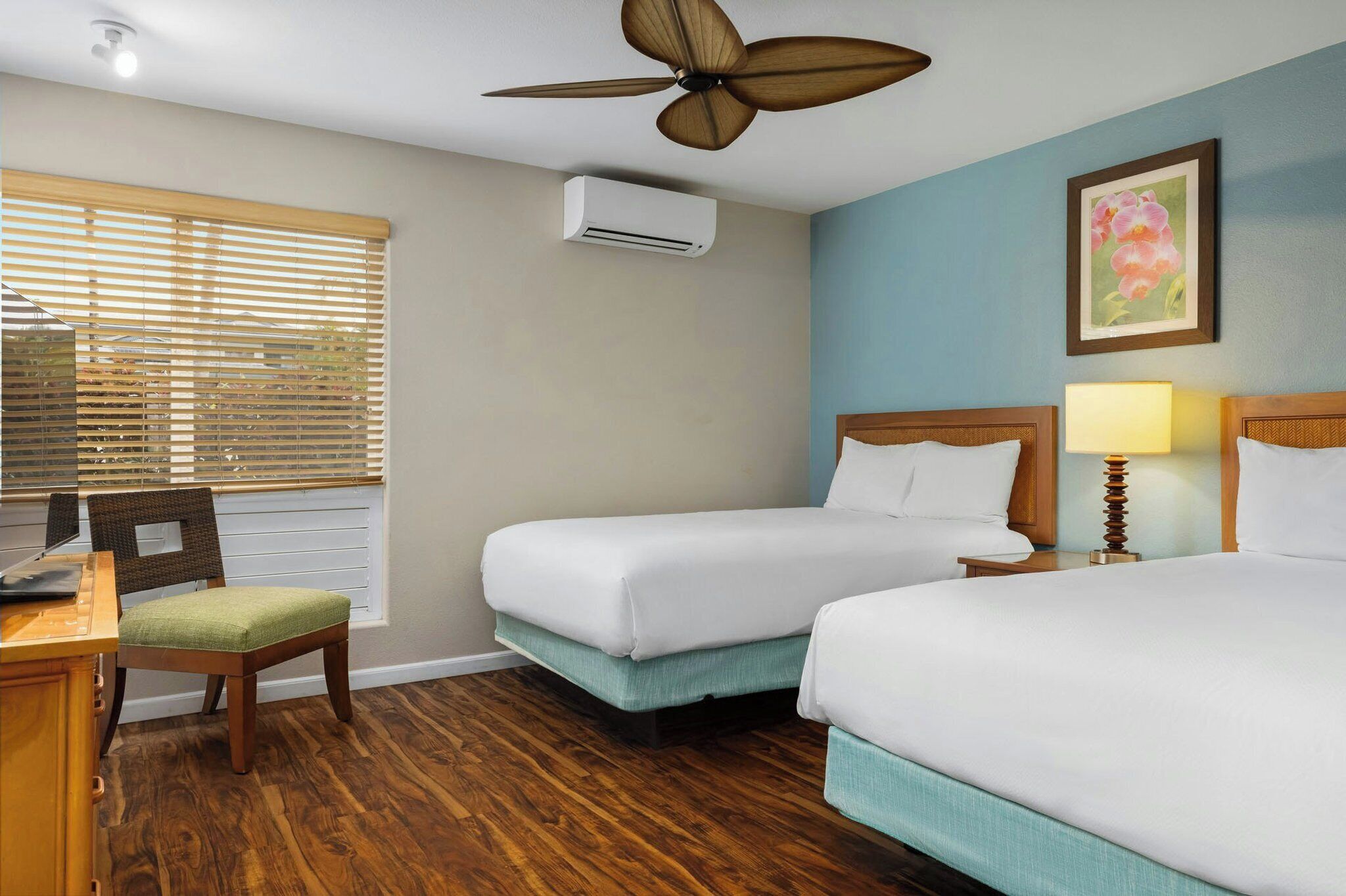 Ein Hotelzimmer mit zwei Betten, einem Schreibtisch mit Stuhl, einem Deckenventilator, einer Klimaanlage und einem Fenster mit Jalousien.