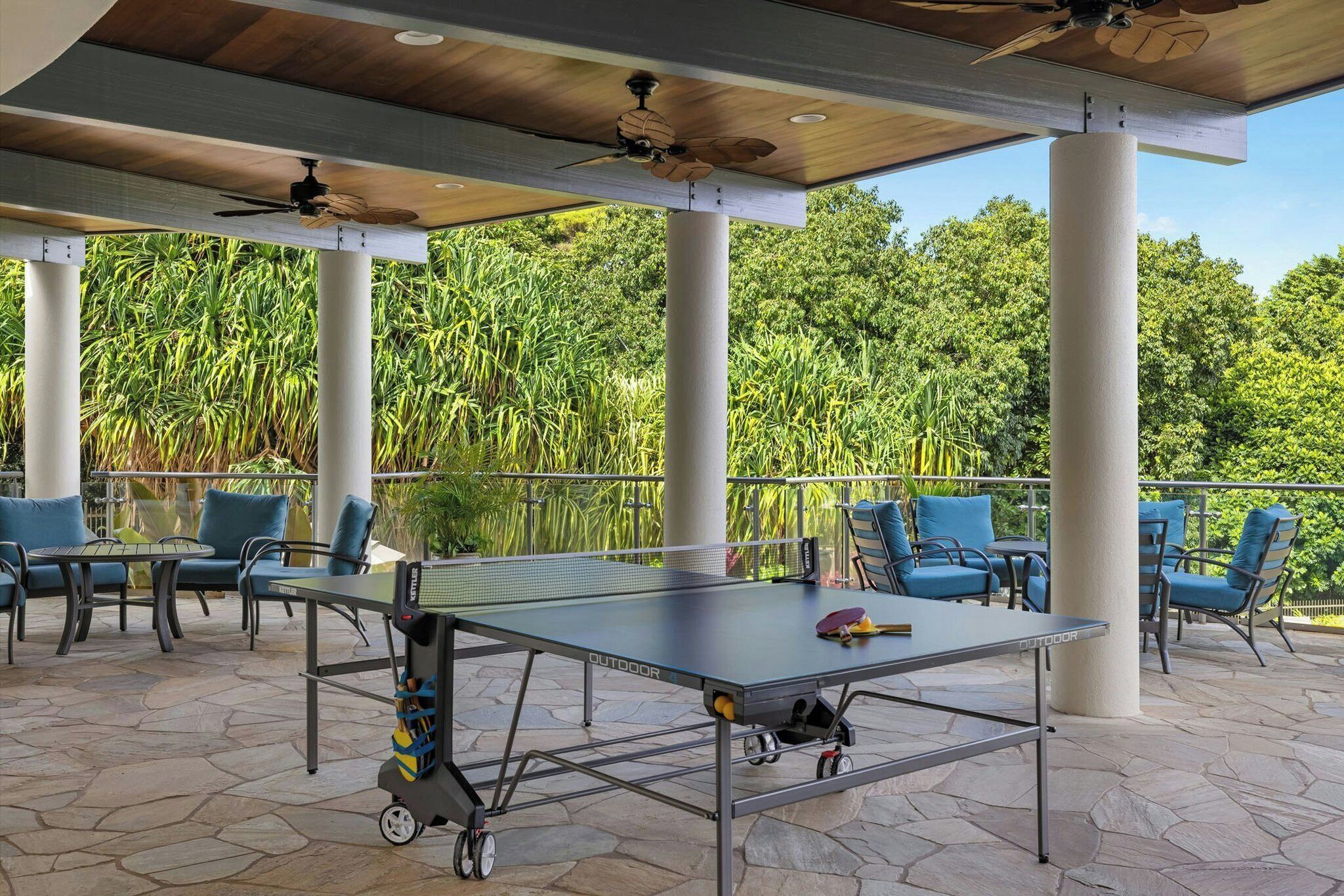 An outdoor table tennis setup with chairs, a ceiling fan, and a view of lush greenery.