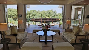 A spacious indoor area with a round table surrounded by cushioned seating, a large window offering a view of lush greenery, and a ceiling light fixture. - Club Wyndham Bali Hai Villas (Princeville)
