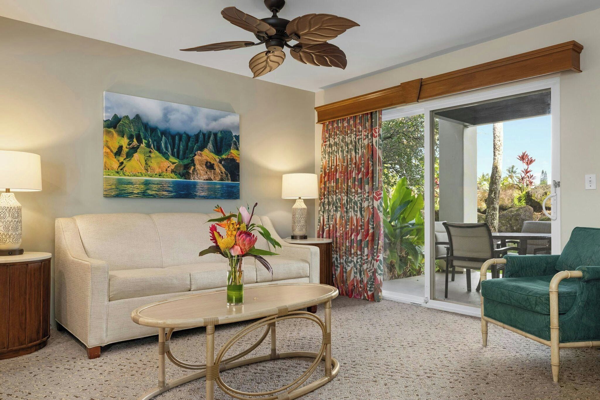 A hotel room with a sofa, a table with a vase of flowers, a ceiling fan, a painting of a landscape, and a view through a glass door.