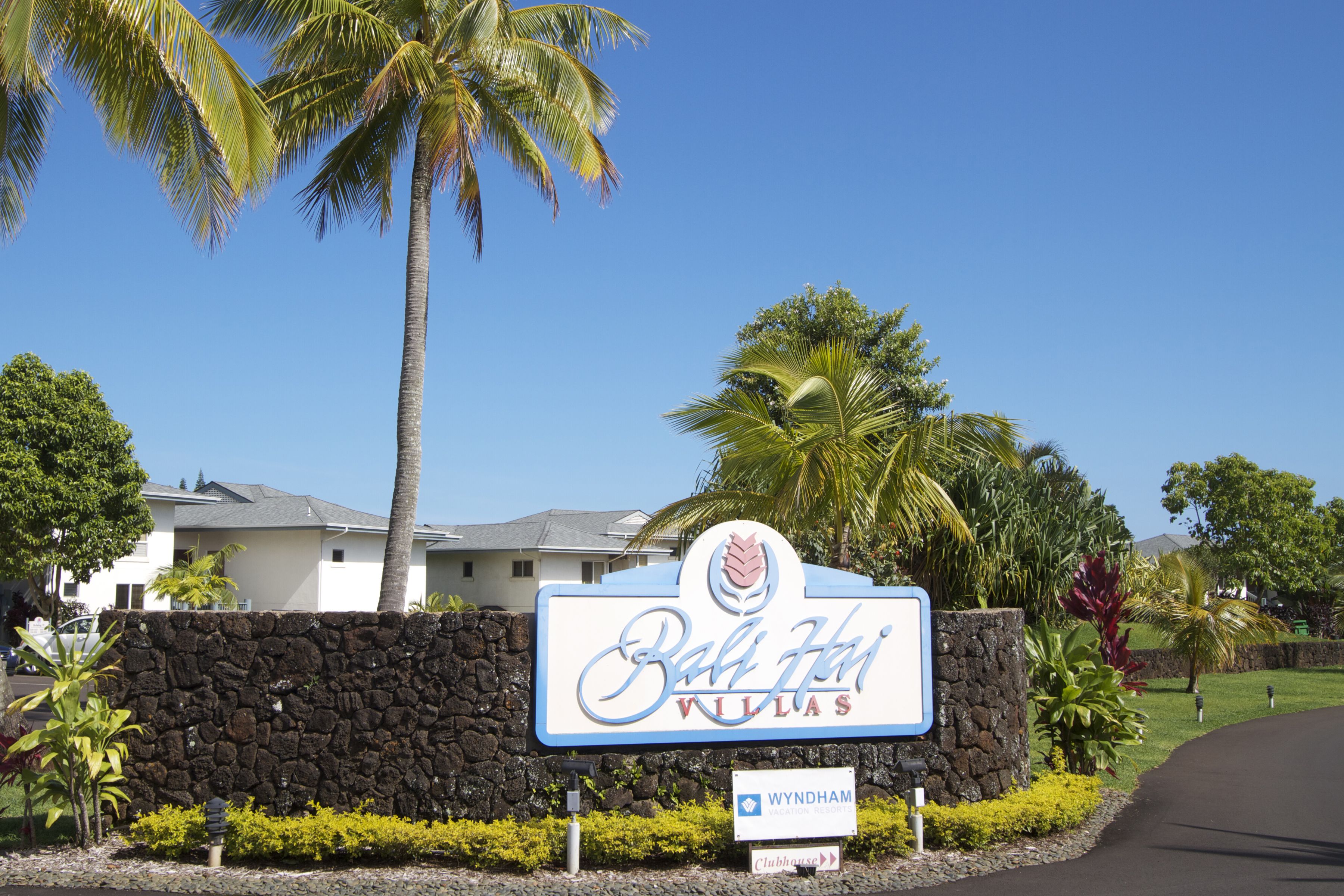 A sign for Bali Hai Villas with a stone wall, palm trees, and a clear blue sky.