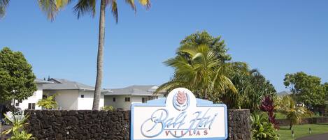 A sign for Bali Hai Villas with a stone wall, palm trees, and a clear blue sky.