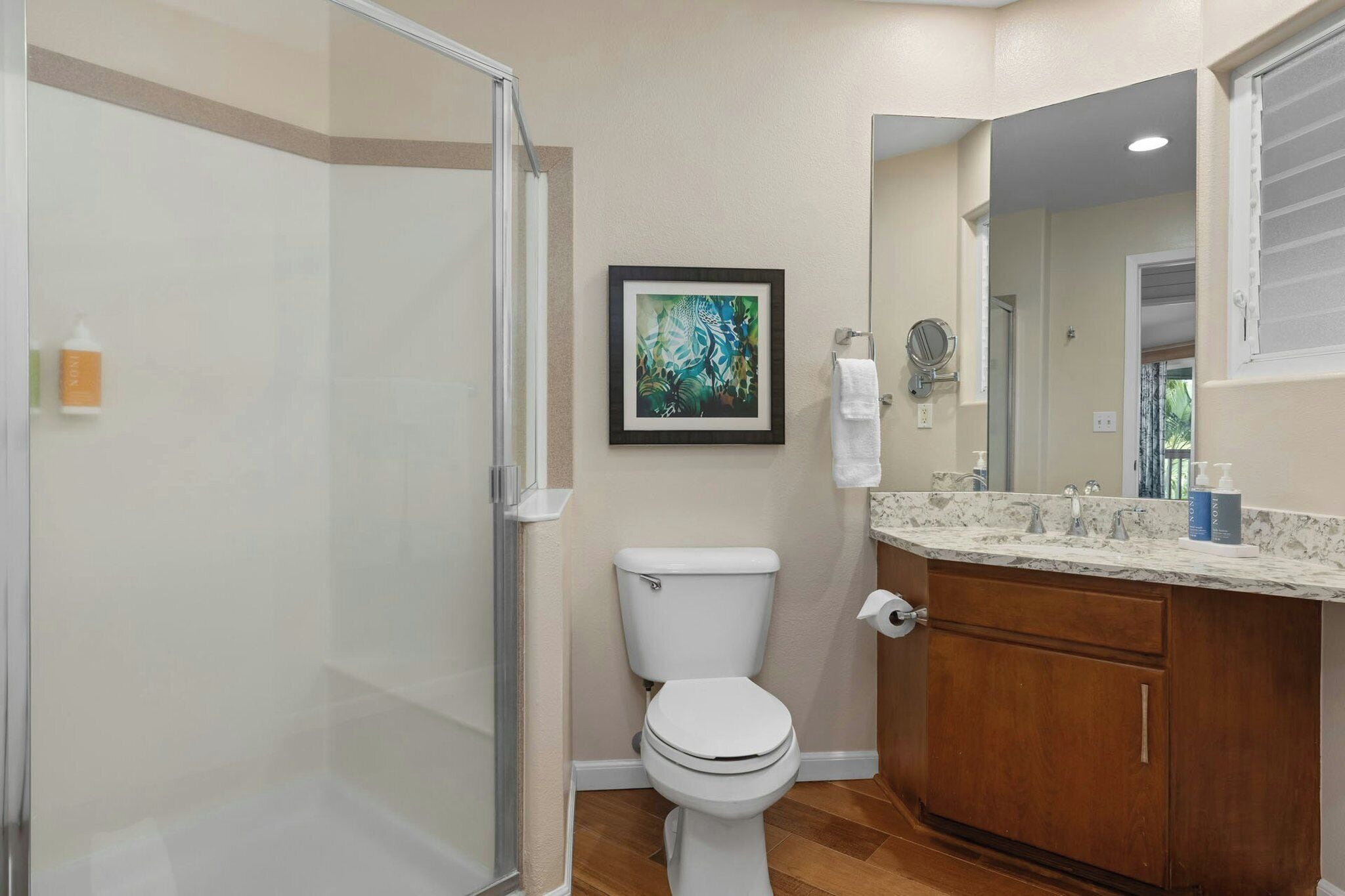 A bathroom with a toilet, a shower enclosure, a framed artwork, a mirror, and a marble countertop.