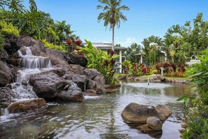 Une cascade d’eau qui dévale des rochers pour se jeter dans un bassin, le tout entouré d’une végétation tropicale luxuriante, avec un bâtiment visible au loin.