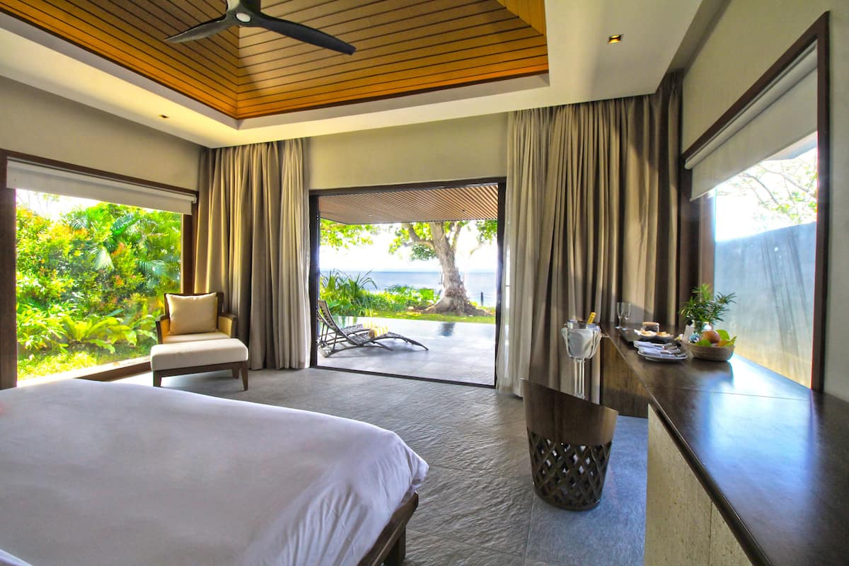 A hotel room with a bed, a chair, a dining table with a view, and a large window with curtains.