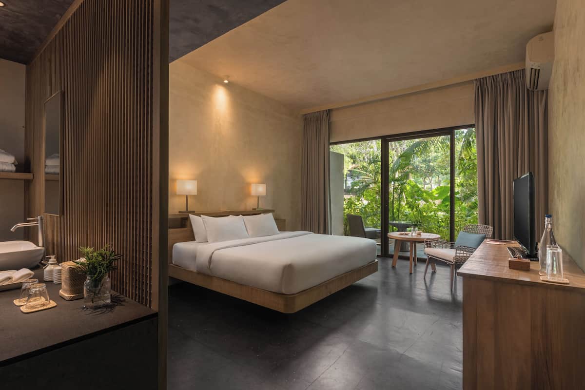 A modern hotel room with a large bed, bedside lamps, a desk with a chair, a TV, and a view of greenery through the window.