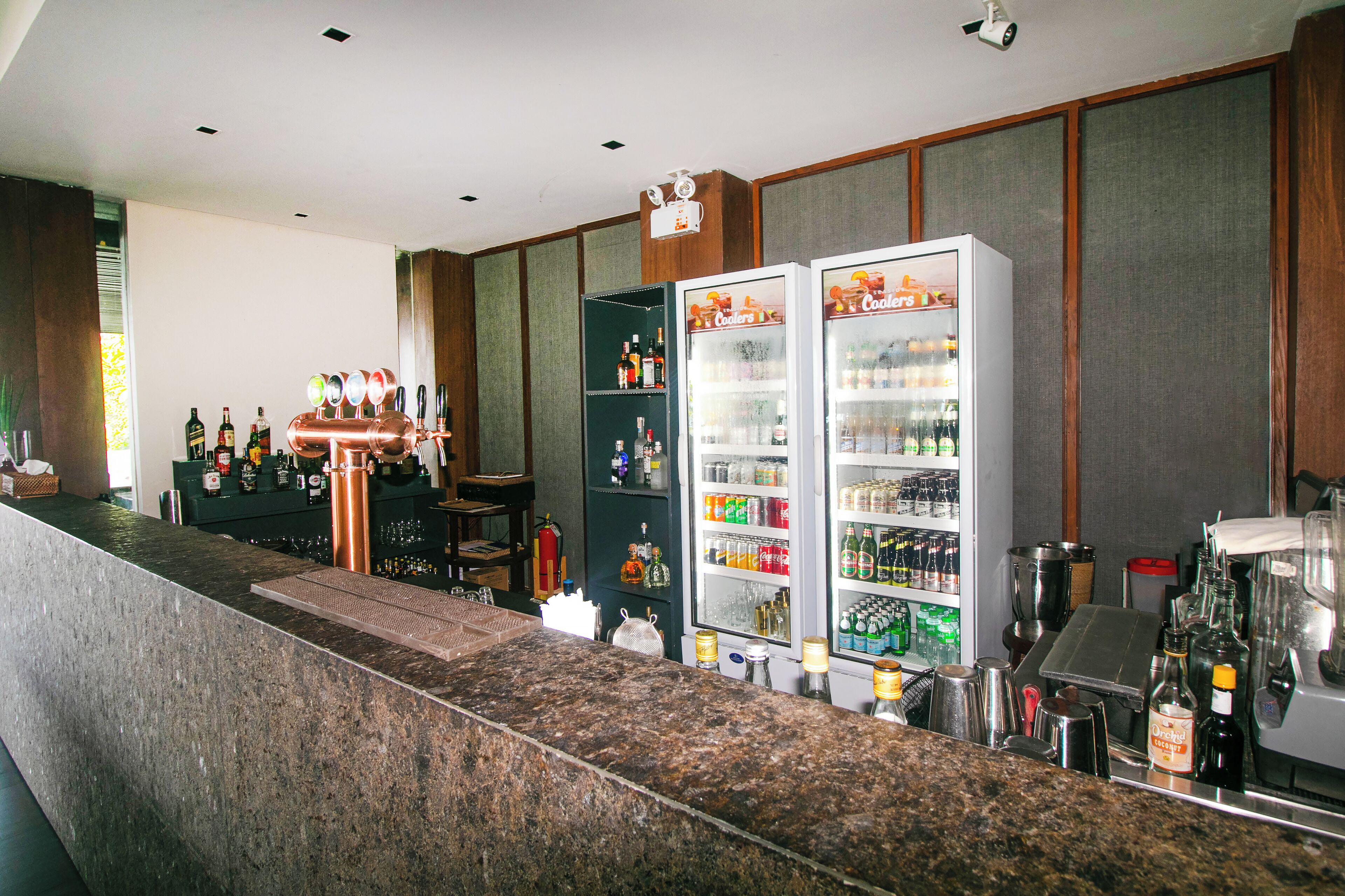 A bar area with a granite countertop, two refrigerators stocked with beverages, and a variety of bottles and glasses on shelves.