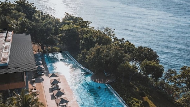 Vista aérea de un resort con una piscina rectangular rodeada de sillas de descanso y sombrillas, con el mar tranquilo y abundante vegetación de fondo.