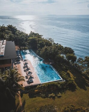 Vista aérea de un resort con una piscina rectangular rodeada de sillas de descanso y sombrillas, con el mar tranquilo y abundante vegetación de fondo.