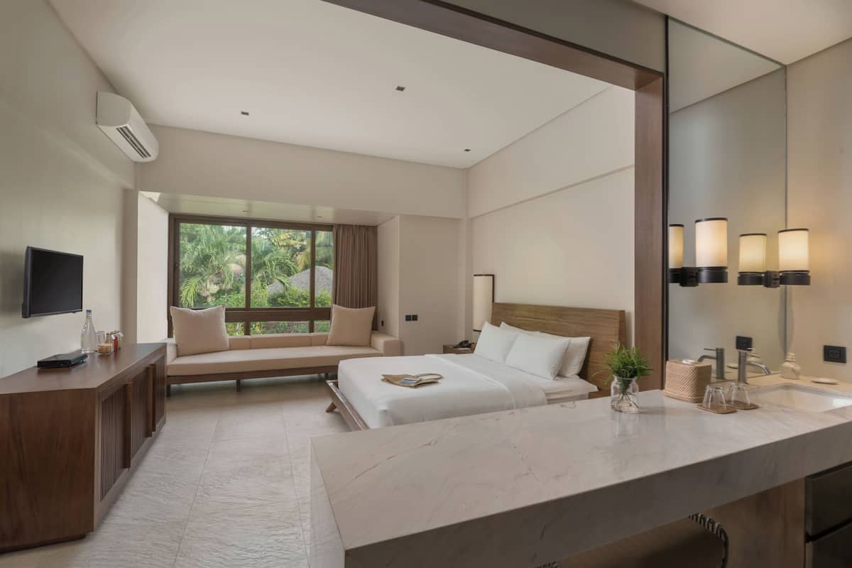 A modern hotel room with a large bed, a desk, a TV, and a large window with a view of greenery.
