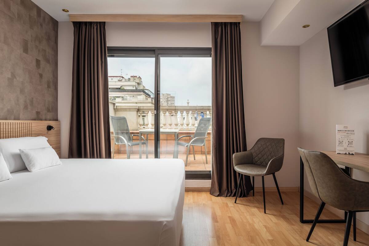 A hotel room with a large bed, a desk with two chairs, a TV mounted on the wall, and a balcony with a table and chairs.