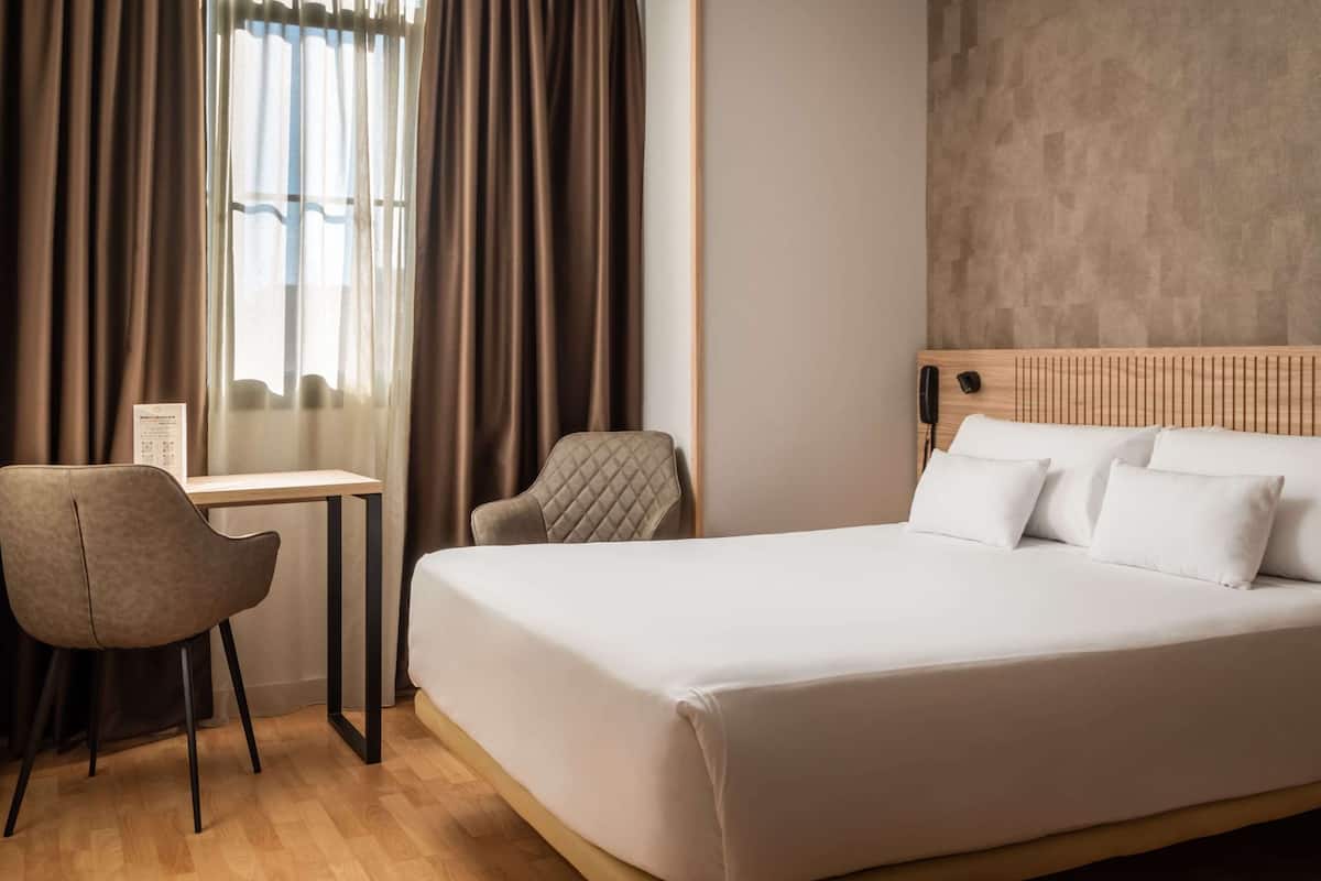 A hotel room with a bed, a desk, a chair, and a window with curtains.