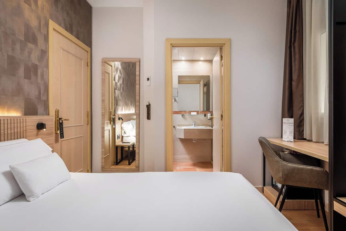 A hotel room with a bed, a desk, a chair, a bathroom with a mirror, and a door leading to another room.