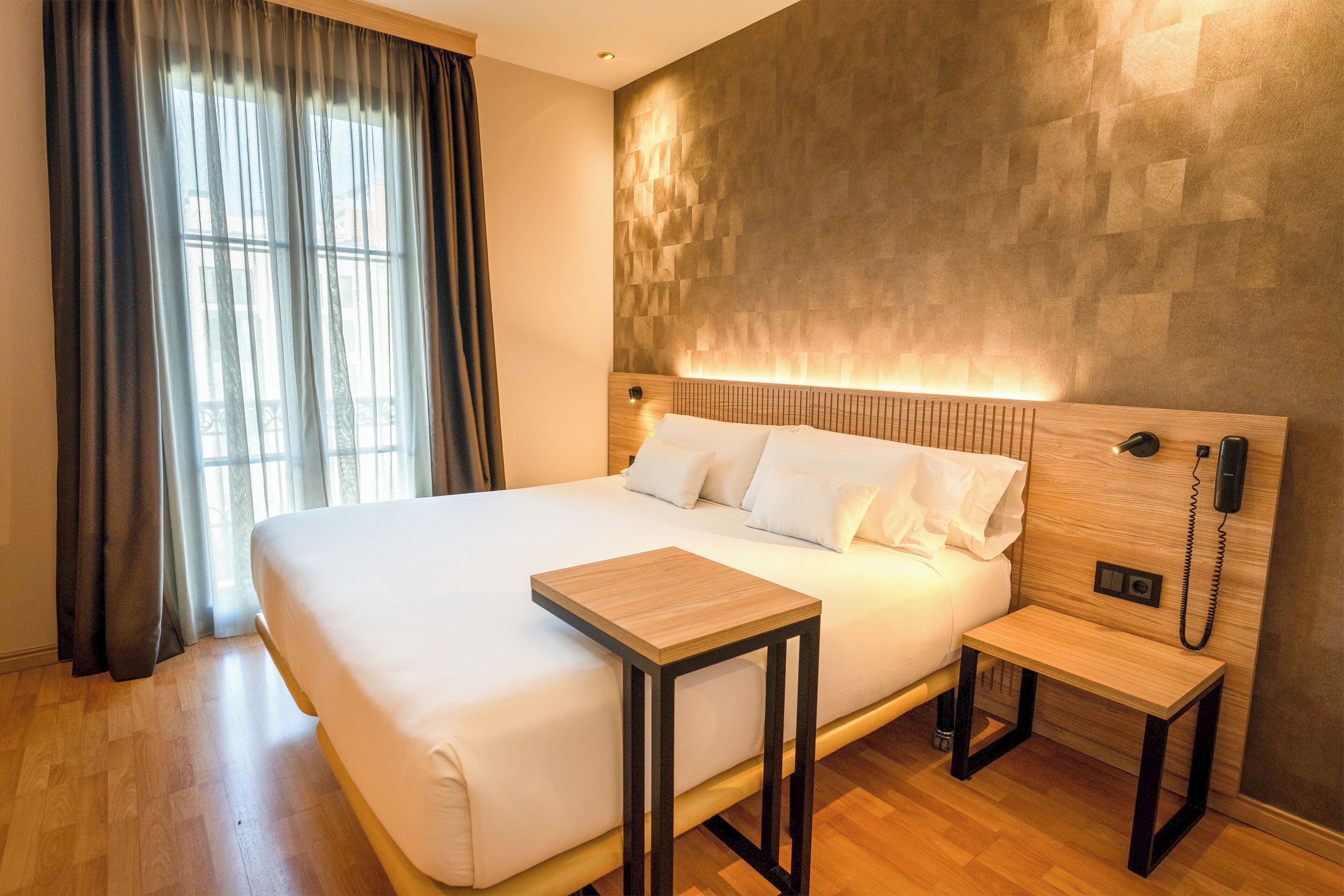 Una habitación de hotel moderna con una cama grande, una mesita de noche, un teléfono y una ventana con cortinas.