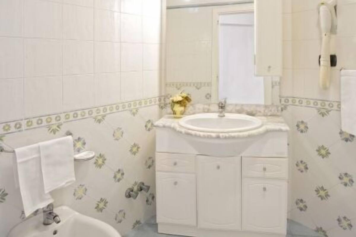 Un baño con lavamanos blanco, espejo y bidé. Las paredes tienen un estampado floral, y hay una alfombra blanca en el suelo.