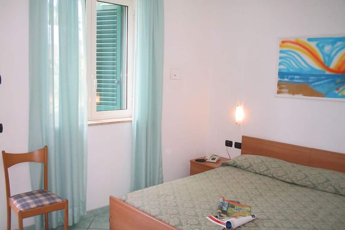 Un dormitorio con cama, una silla, ventana con persianas, mesita de noche con lámpara y un cuadro en la pared.