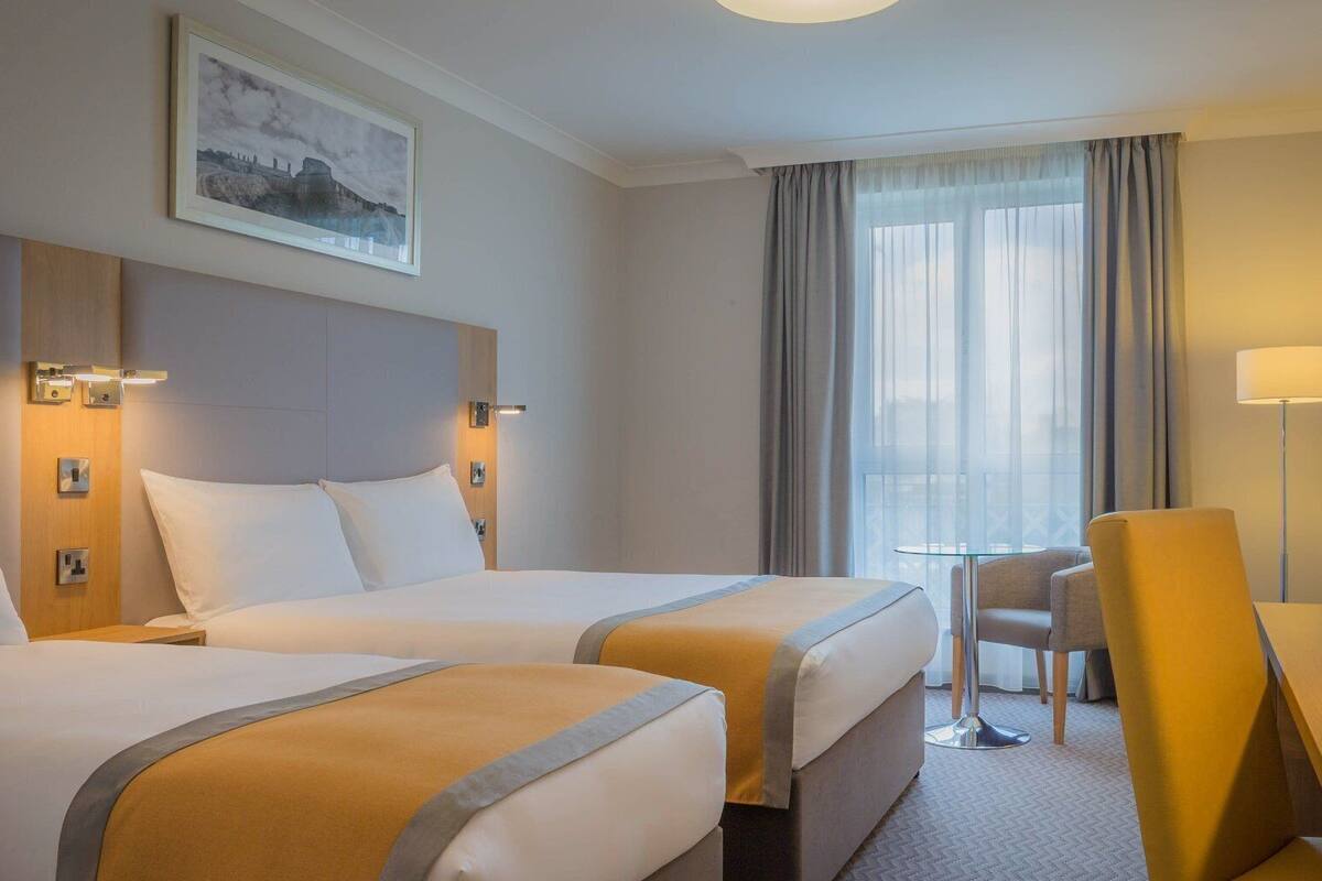Habitación de hotel con dos camas, un escritorio, una silla, una mesita y una ventana con cortinas.